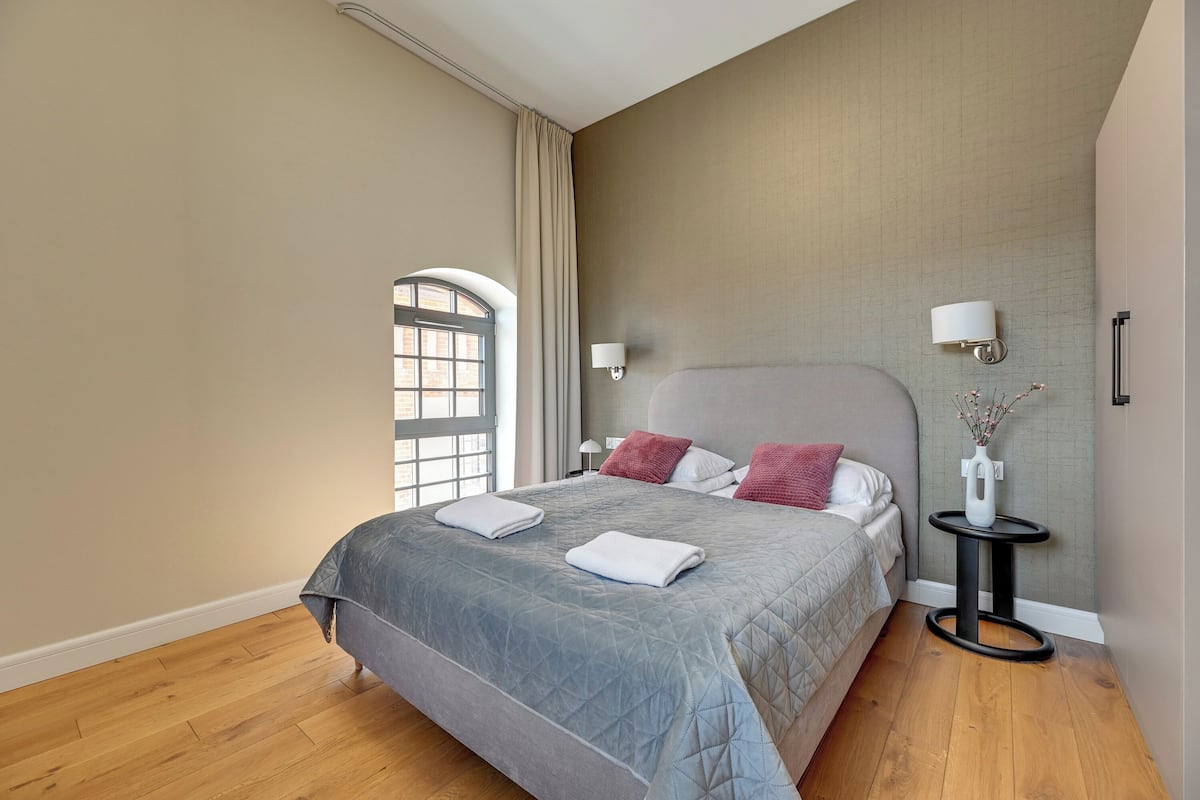 Ein modernes Schlafzimmer mit einem großen Bett, einem Nachttisch, einer Vase mit Blumen und einem Fenster mit Vorhängen.