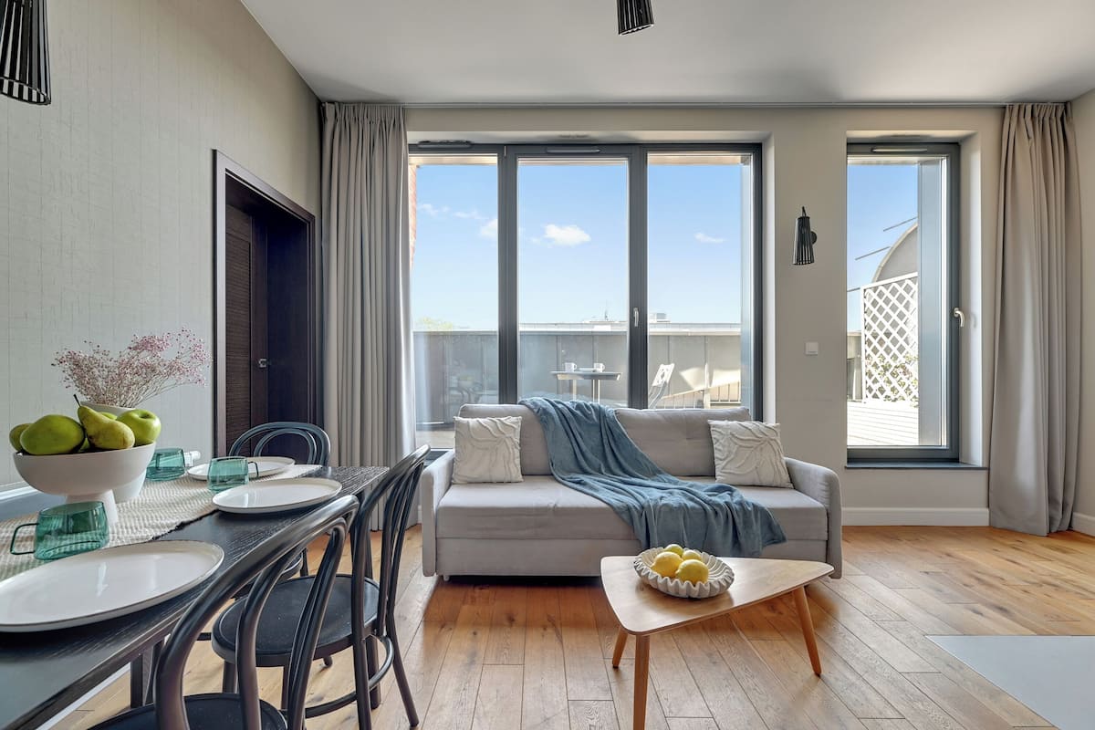 Ein modernes Wohnzimmer mit Sofa, Esstisch und Stühlen. Große Fenster mit Vorhängen, ein Balkon und Blick zum Himmel.