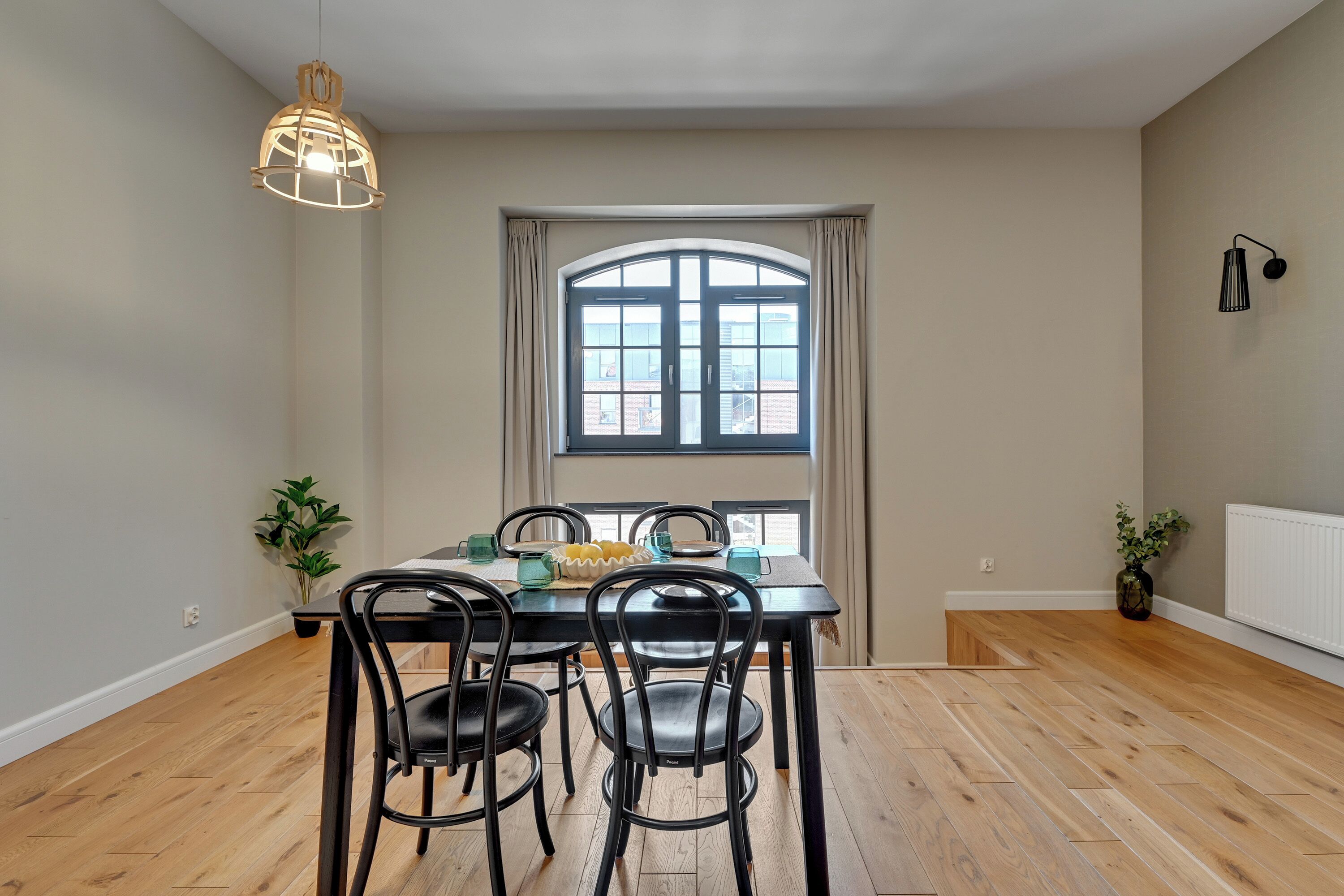 Comedor con una mesa dispuesta para cuatro personas, un ventanal con cortinas, un colgante de luz y una planta en maceta.