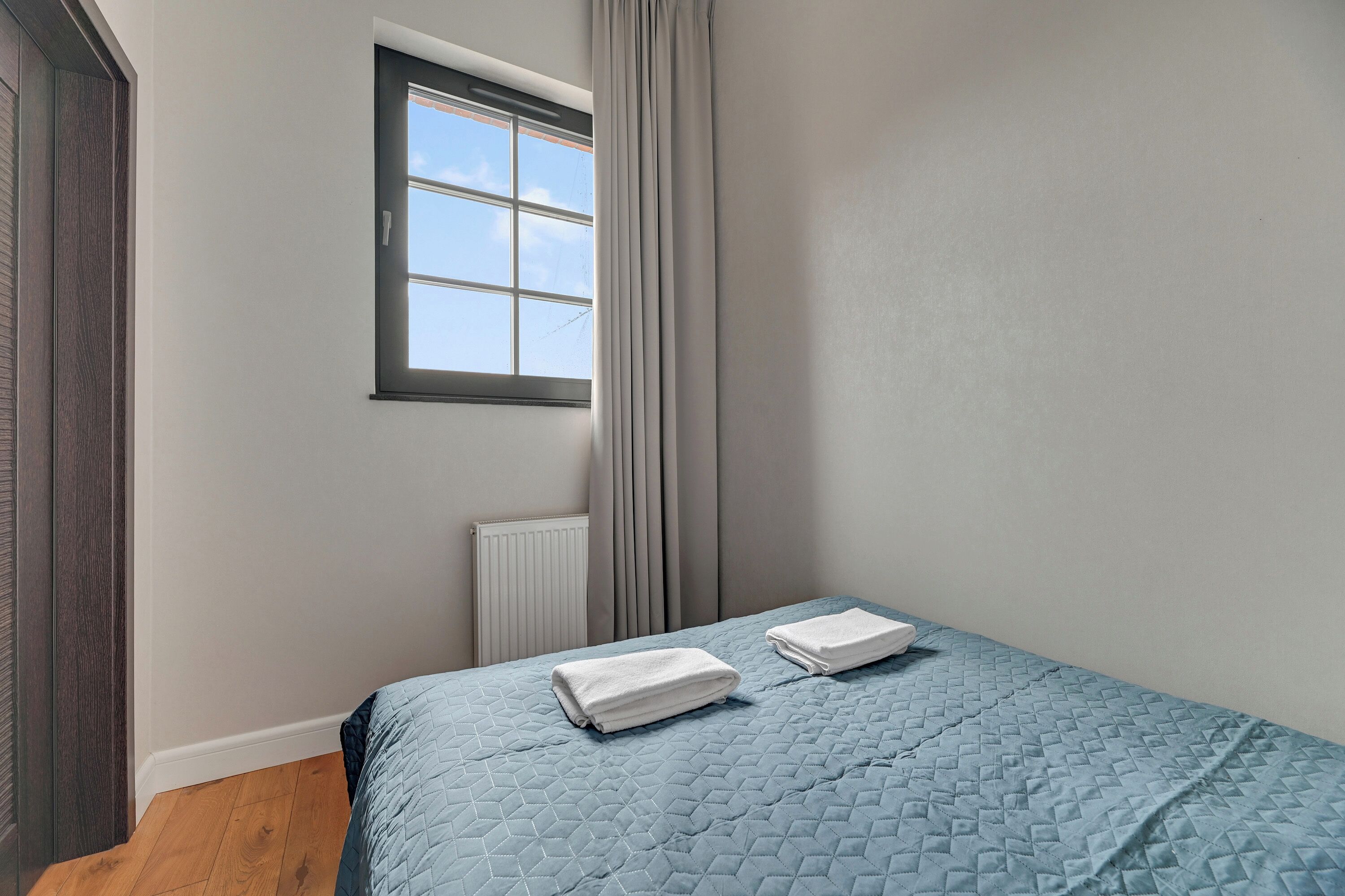 A bedroom with a bed, two neatly folded white towels, a window with curtains, and a radiator.