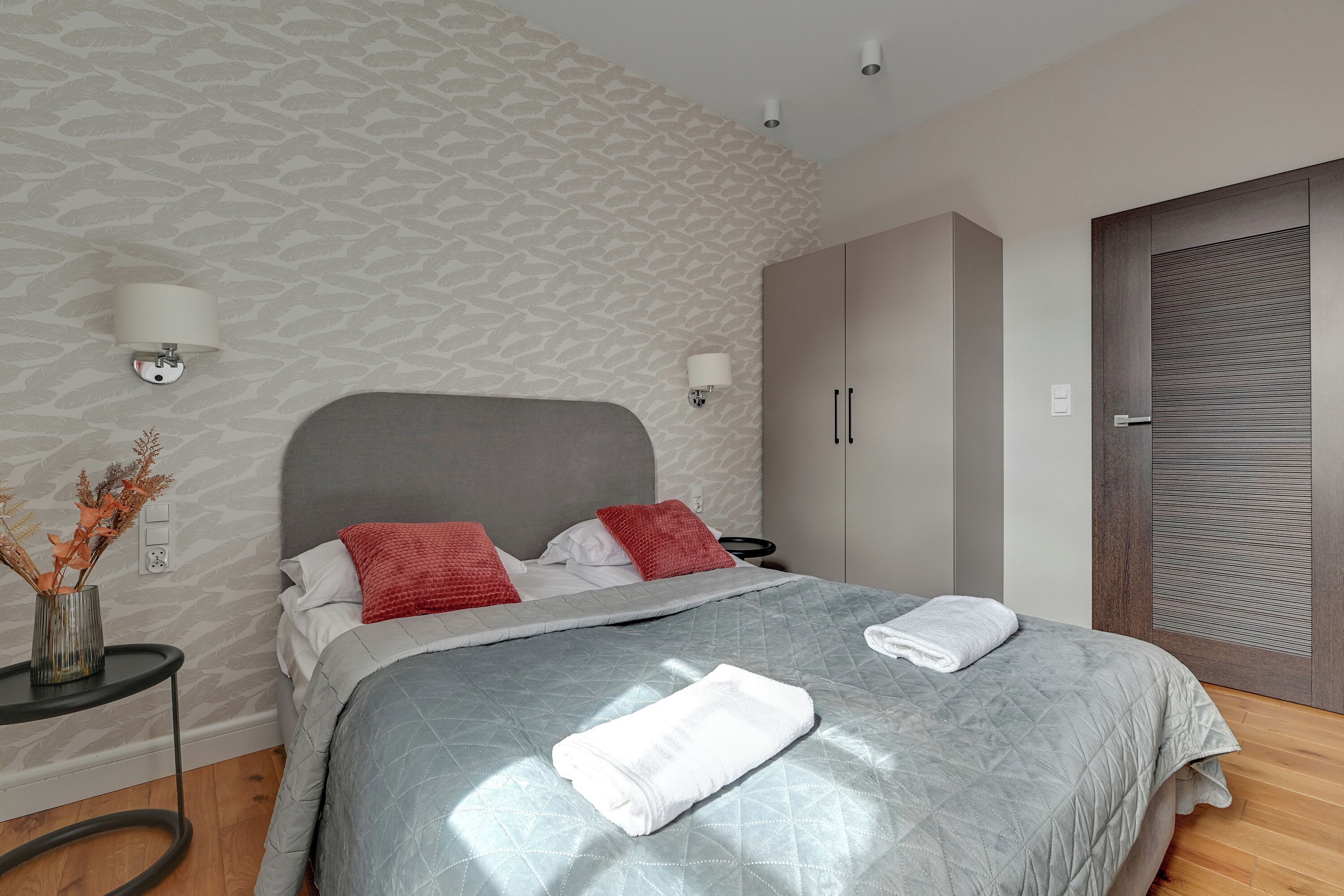 A neatly made bed with a grey quilt, two red pillows, and a white towel.