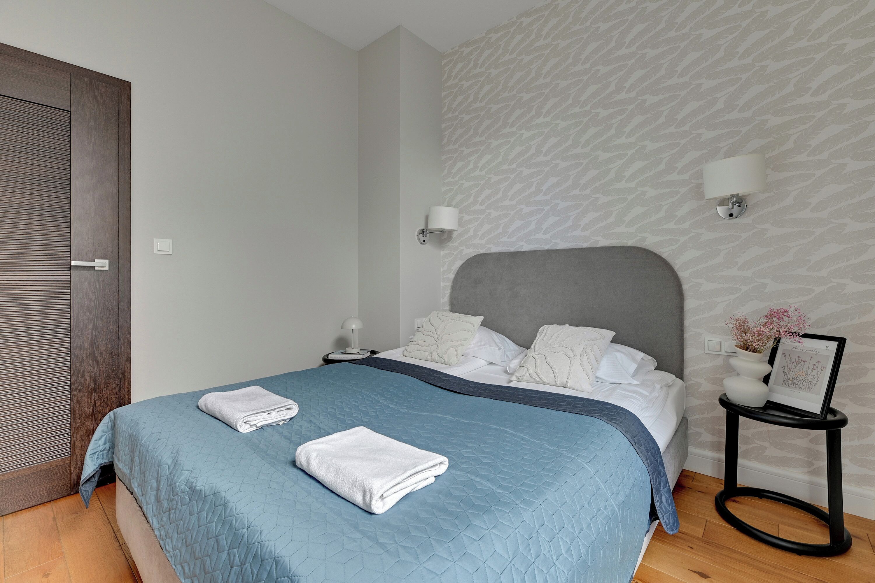 A neatly made bed with a blue quilt, white pillows, and a grey headboard. There are two bedside tables, one with a lamp and the other with a framed picture. The room has a patterned wallpaper and a wooden floor.