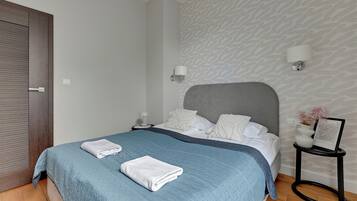 A neatly made bed with a blue quilt, white pillows, and a grey headboard. There are two bedside tables, one with a lamp and the other with a framed picture. The room has a patterned wallpaper and a wooden floor.