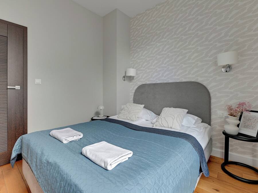 A neatly made bed with a blue quilt, white pillows, and a grey headboard. There are two bedside tables, one with a lamp and the other with a framed picture. The room has a patterned wallpaper and a wooden floor.