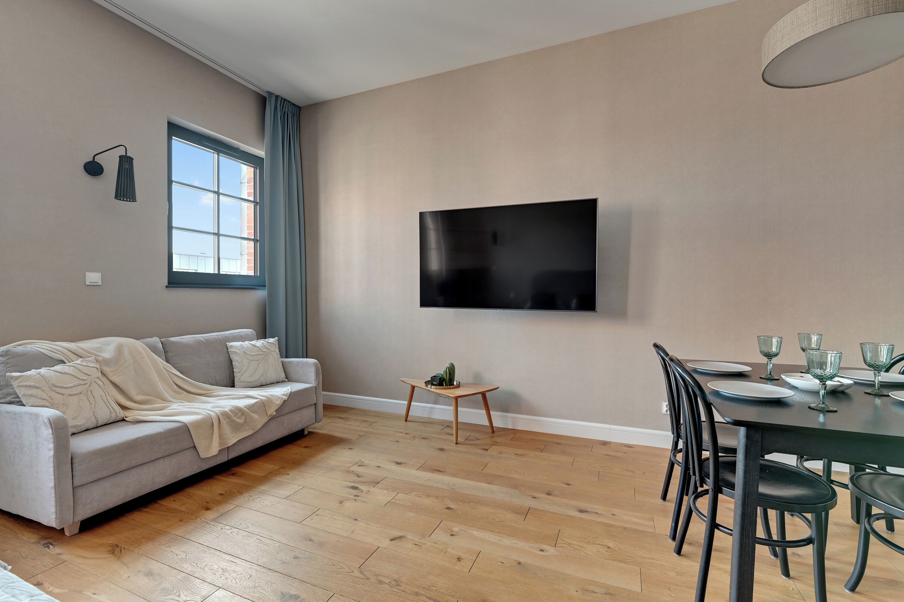 Una sala de estar moderna con un sofá gris, piso de madera, una mesa de comedor para cuatro personas y un televisor de pantalla plana montado en la pared.