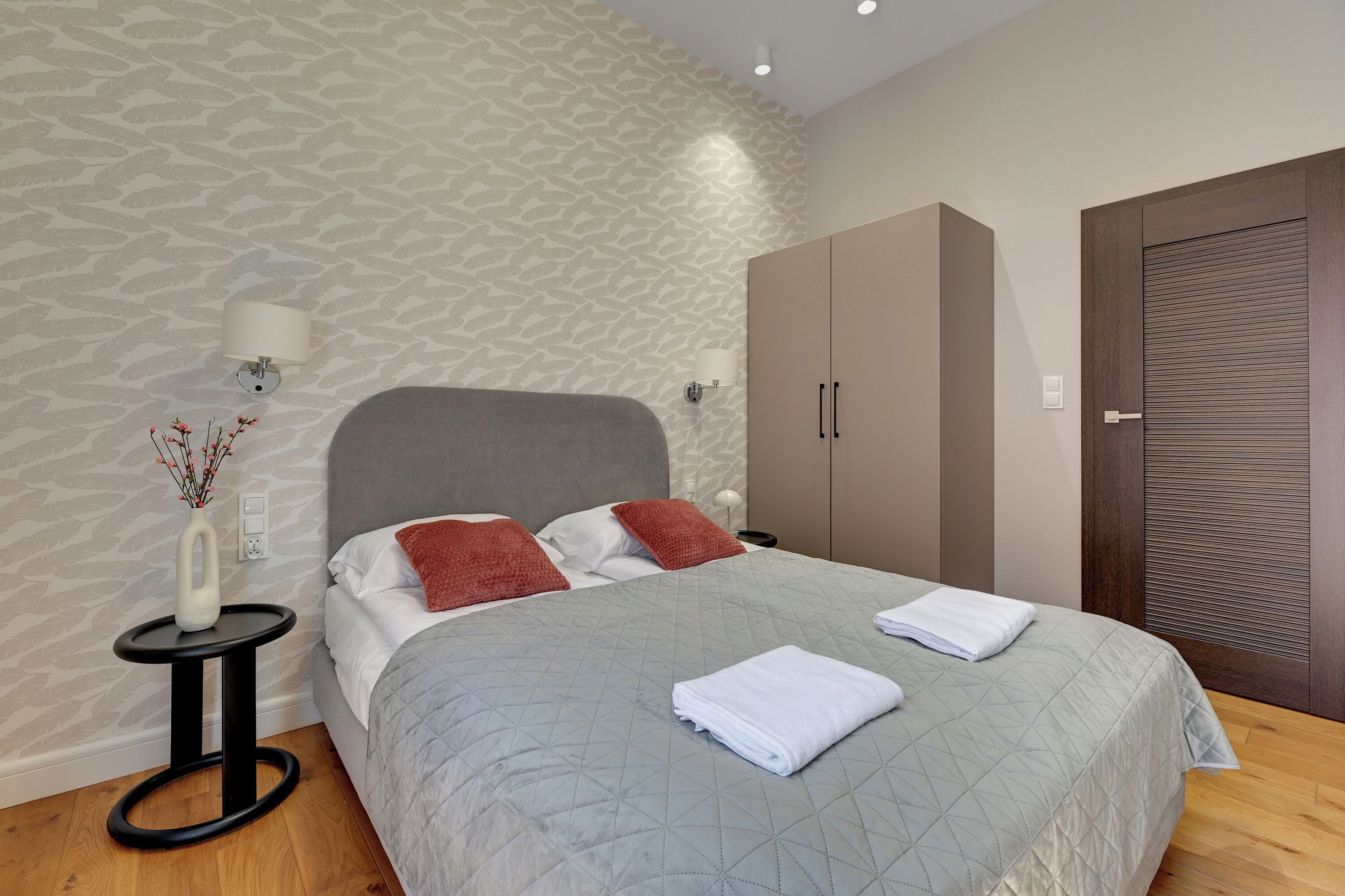 Un letto rifatto con una coperta grigia, un comodino con un vaso e un armadio con sportello scuro.