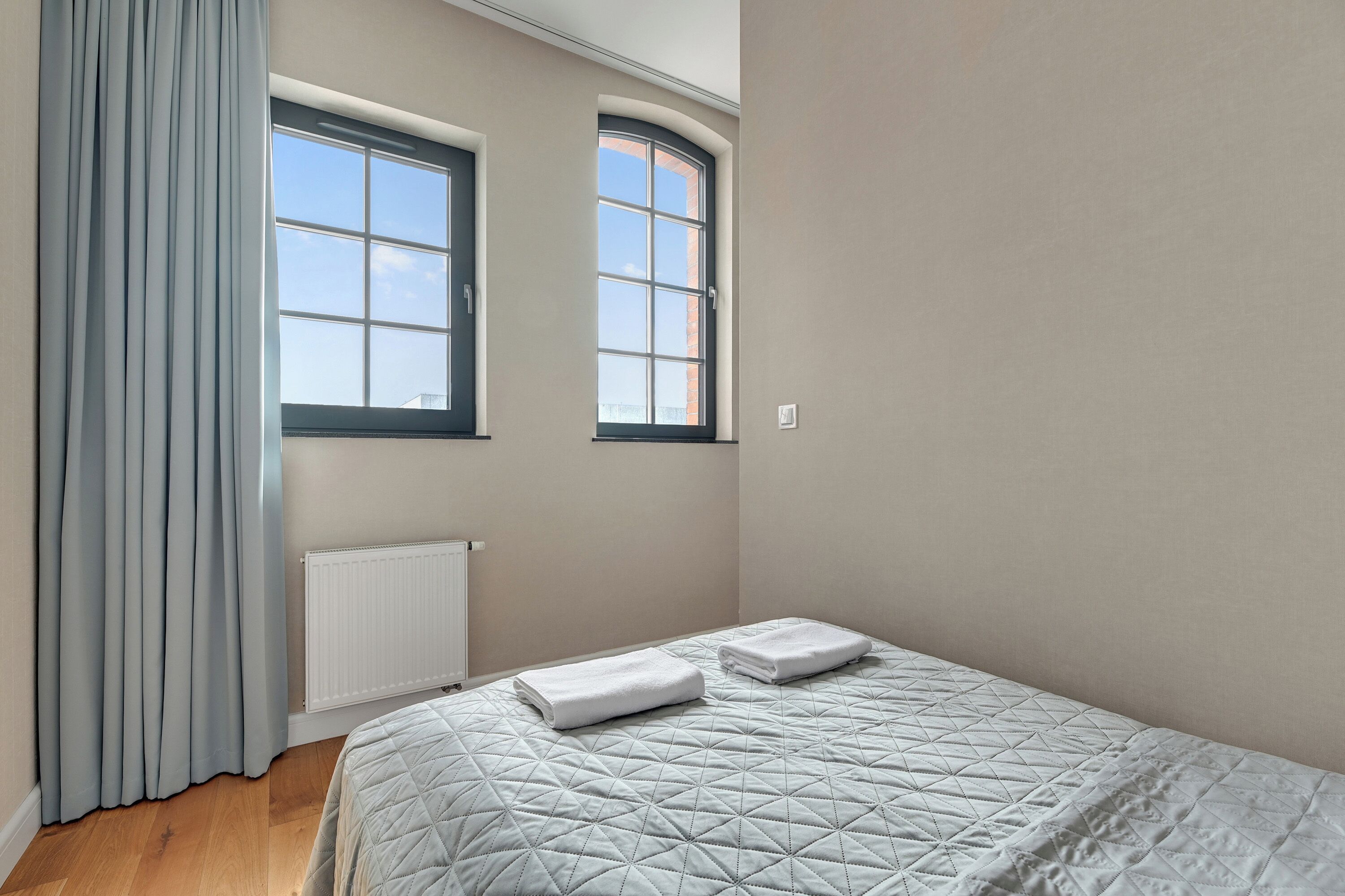Un dormitorio con una cama, dos ventanas, un radiador y una cortina.
