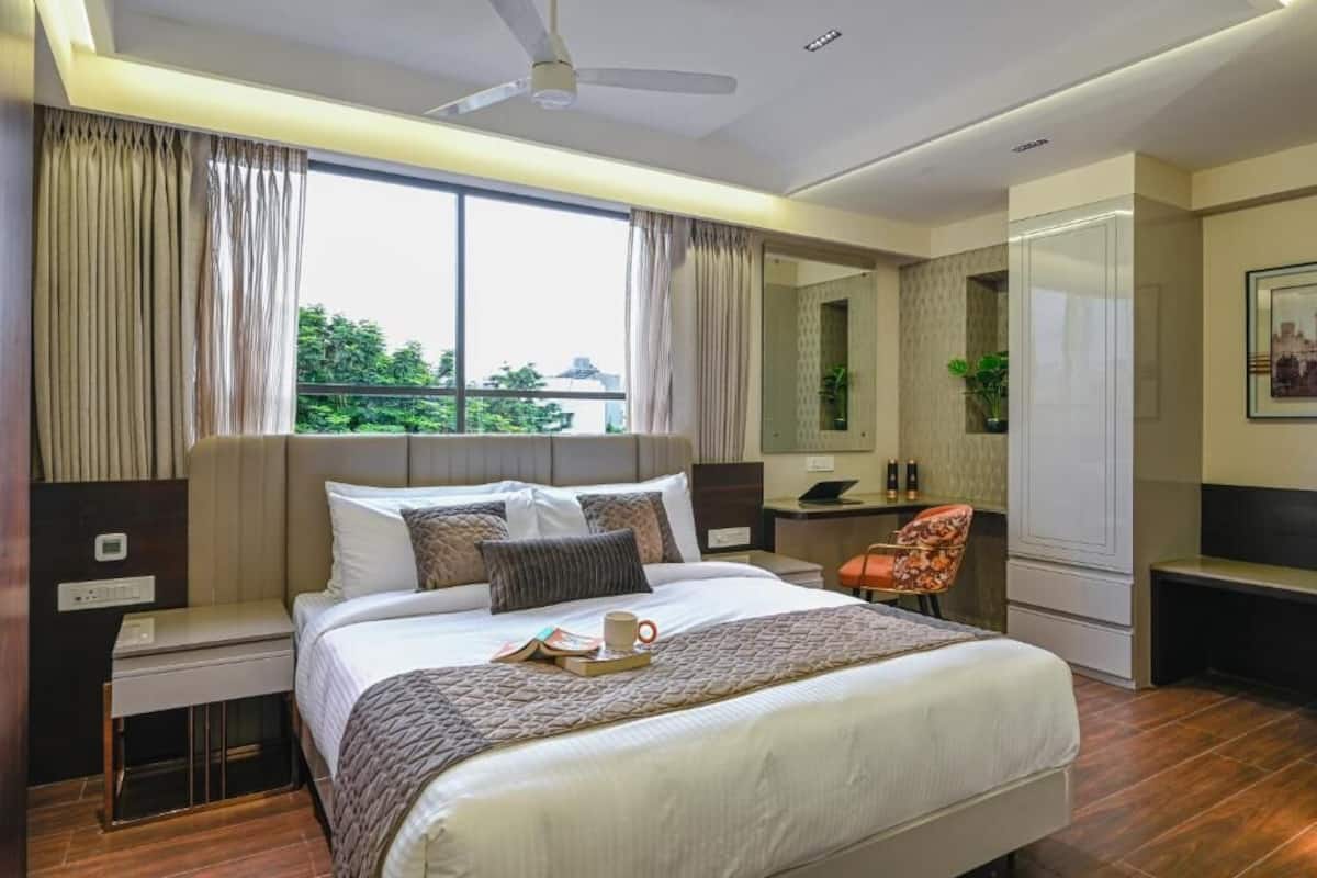 A modern hotel room with a large bed, a desk, a chair, and a view of greenery outside.