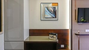 A modern interior with a wooden bench, a framed abstract painting, and a television on a wall. - Hotel Grand Girdhar Premier (Indore)