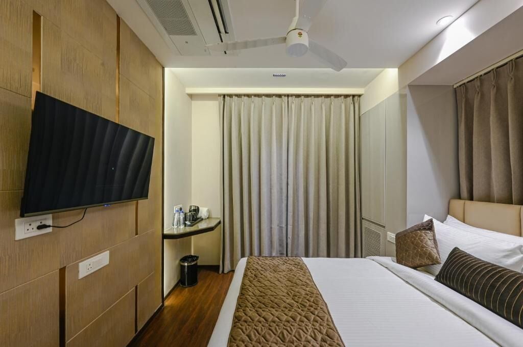 Hotelkamer met een bed, een aan de muur bevestigde televisie, een plafondventilator, een klein tafeltje met een kopje en een prullenbak, en een raam met gordijnen.