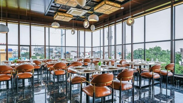A modern restaurant with a high ceiling, large windows, and a view of the cityscape. Tables are set with chairs, and the floor is polished. - Hotel Grand Girdhar Premier (Indore)