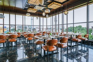 A modern restaurant with a high ceiling, large windows, and a view of the cityscape. Tables are set with chairs, and the floor is polished. - Hotel Grand Girdhar Premier (Indore)