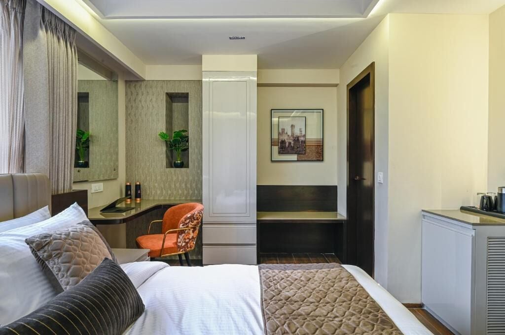 Une chambre d’hôtel avec un lit, un bureau, une chaise, un miroir, une plante et un tableau encadré au mur.