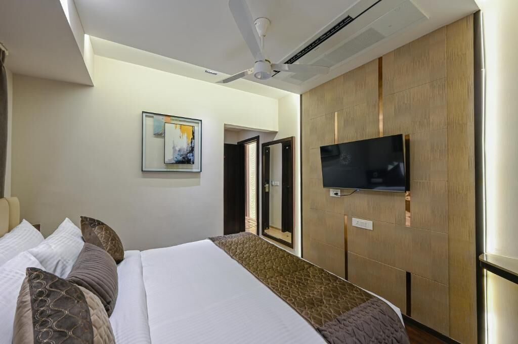 A hotel room with a bed, a flat-screen TV mounted on the wall, a ceiling fan, and a painting on the wall.