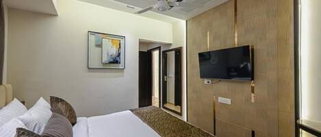 A hotel room with a bed, a flat-screen TV mounted on the wall, a ceiling fan, and a painting on the wall.