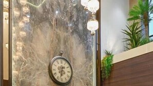 A lobby with a marble wall, a clock, two orange chairs, a small table with a plant, and a tall floor lamp. - Hotel Grand Girdhar Premier (Indore)