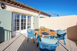 Um pátio externo com uma mesa redonda de madeira posta para quatro pessoas, rodeado por cadeiras de vime azul com almofadas.