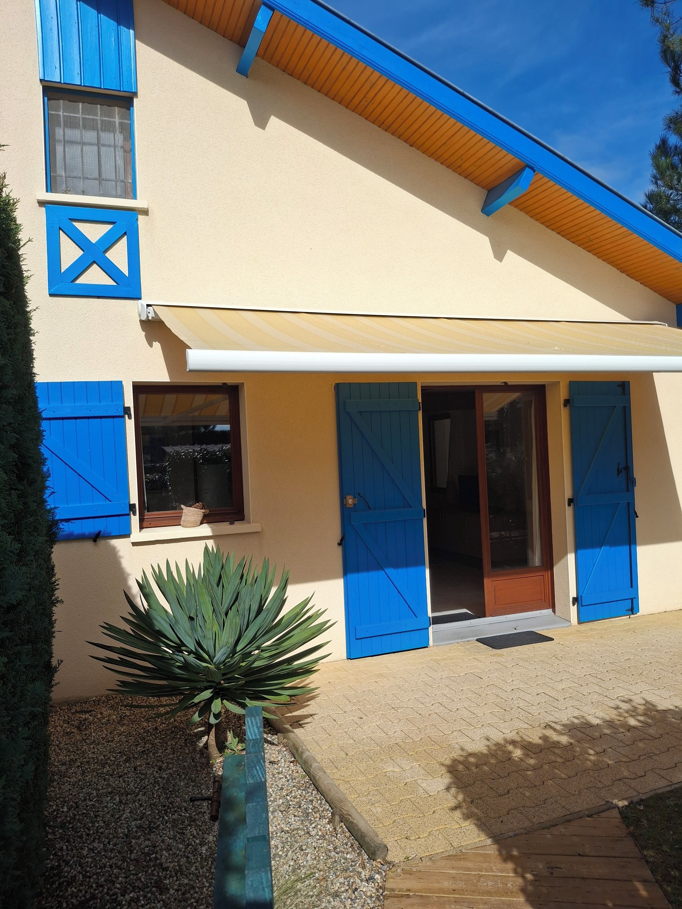 Une maison à deux étages avec des volets bleus, une porte en bois et une petite fenêtre ornée d’une plante.
