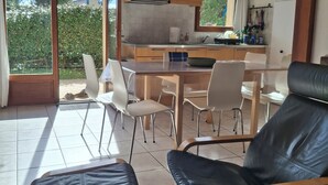 A spacious living area with a dining table, chairs, and a sofa. There is a kitchen area with a microwave and a sink. The room has large windows and wooden beams on the ceiling.