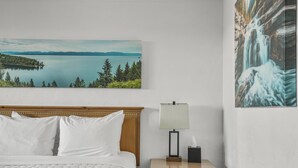 A hotel room with a bed, a nightstand with a lamp, a painting of a lake and forest, and a wall-mounted picture of a waterfall. - Premium Room, 1 Queen Bed, Lake View (Polson)