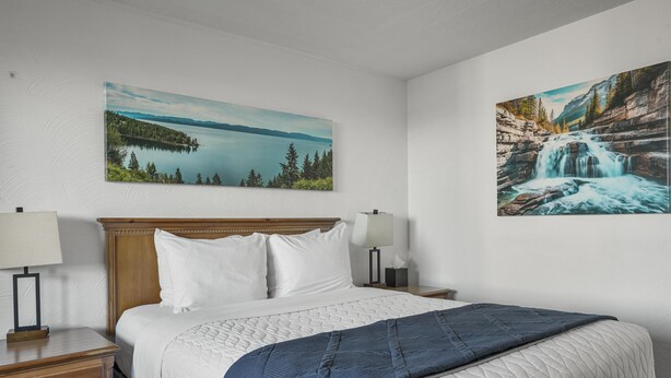 A hotel room with a bed, two bedside tables, a nightstand, a blue blanket, and two paintings on the wall. - Premium Room, 1 Queen Bed, Lake View (Polson)