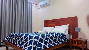 Cama bem arrumada com colcha azul e branca estampada, cabeceira de madeira e mesa de cabeceira com abajur.