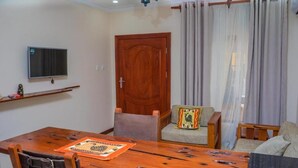 A dining area with a wooden table, chairs, a television on the wall, a door, and a window with curtains. - Upland Serviced Apartments (Lusaka)