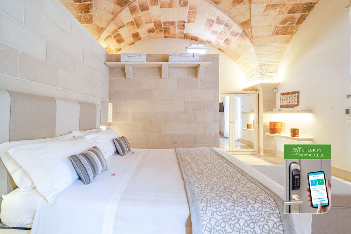 A hotel room with a stone arch ceiling, a bed with white linens, a patterned throw, and a self-check-in kiosk.