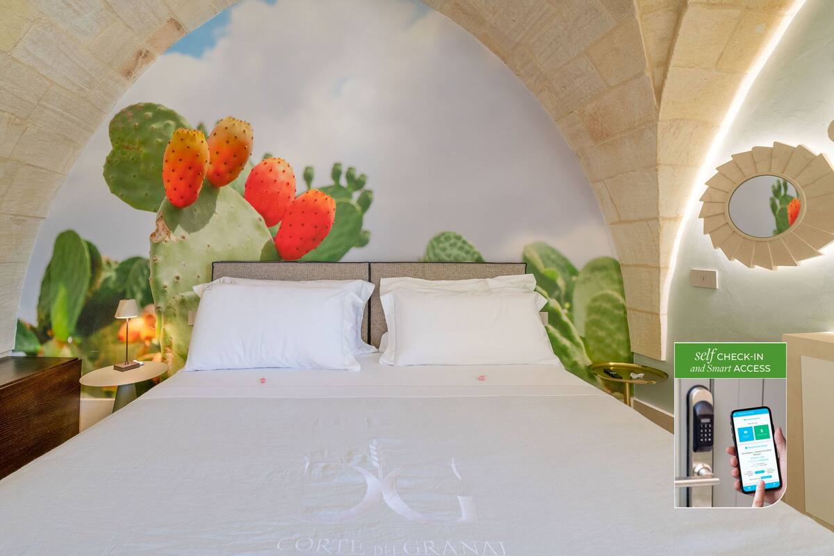 A hotel room with a bed, white pillows, a nightstand with a lamp, a wall mural of cacti, and a self-check-in kiosk.