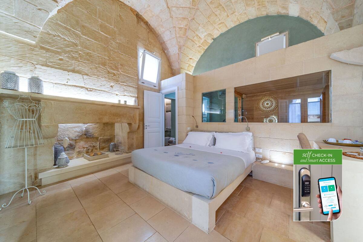 A spacious bedroom with a large bed, a stone wall, a window, and a mirror.