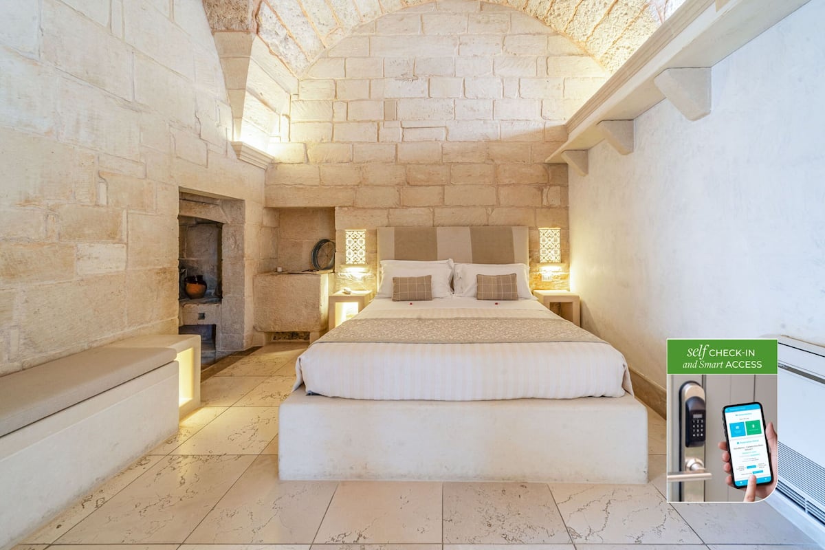 A hotel room with a stone wall, a large bed, a nightstand, and a self-check-in kiosk.