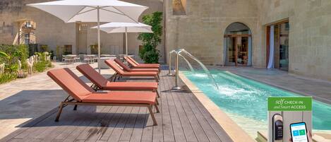 Zona de piscina con sillones reclinables, sombrillas y un quiosco de auto-registro.