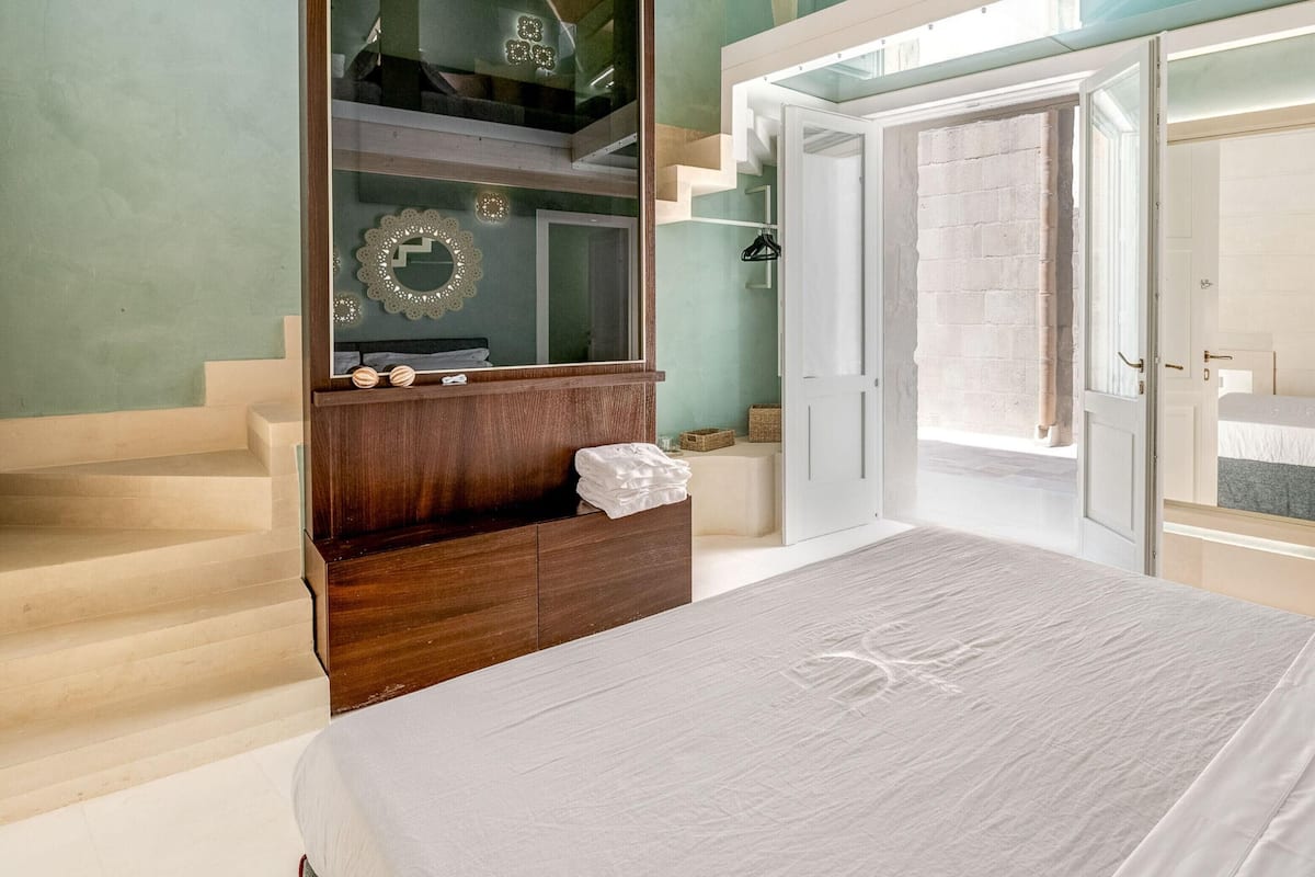 A bedroom with a bed, a large mirror, a wooden dresser, and a door leading to a balcony.
