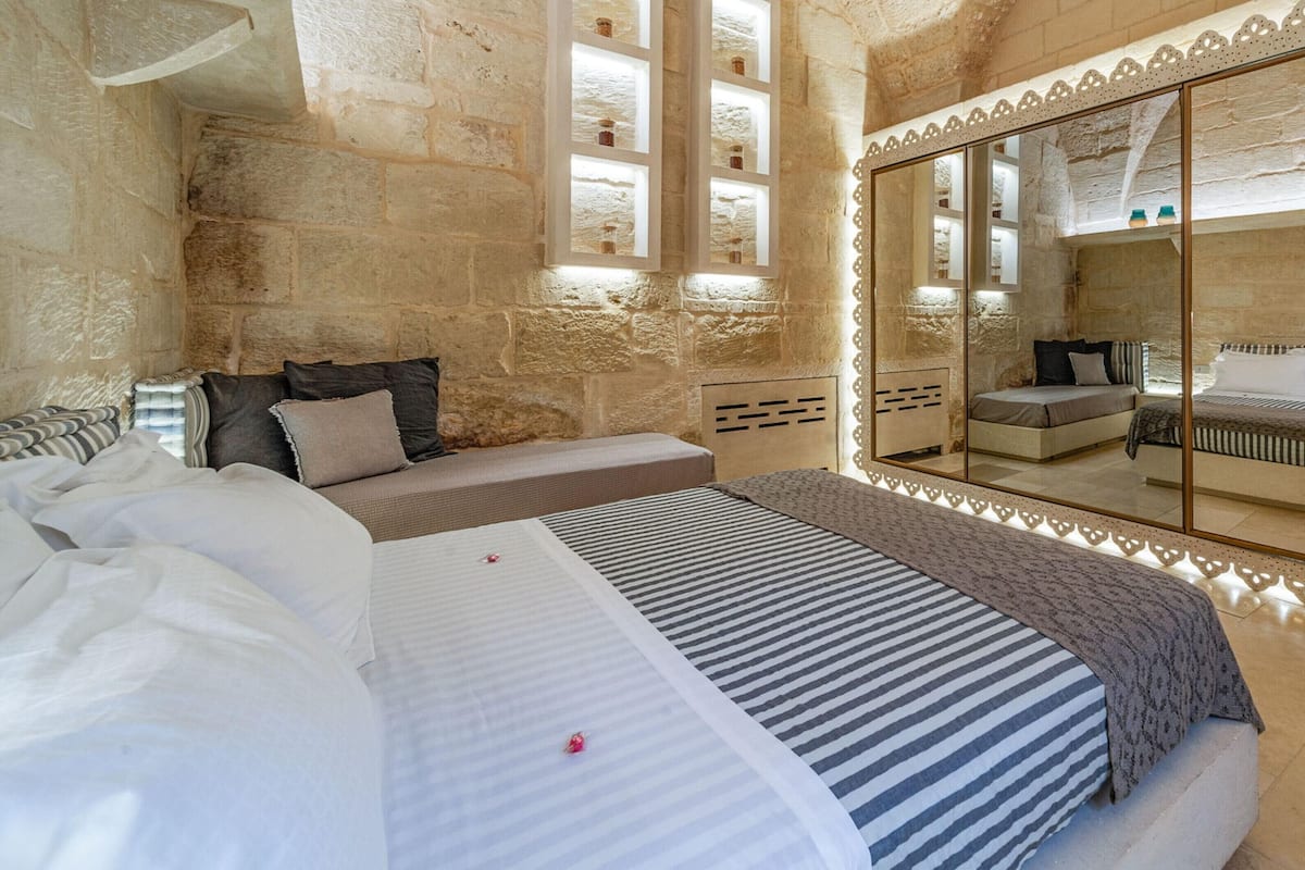 A bedroom with stone walls, a large bed with striped bedding, a bedside table, and a mirror with a decorative frame.