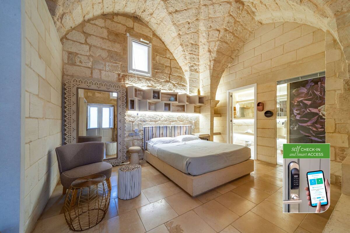 A bedroom with a stone arch ceiling, a bed, a chair, a small table, and a self-check-in kiosk.