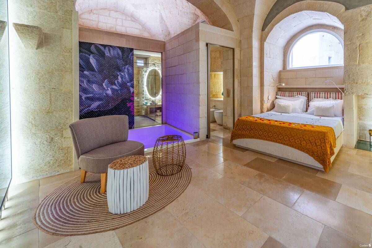 A spacious room with a large bed, a seating area with a chair and ottoman, and a bathroom with a large mirror and sink.