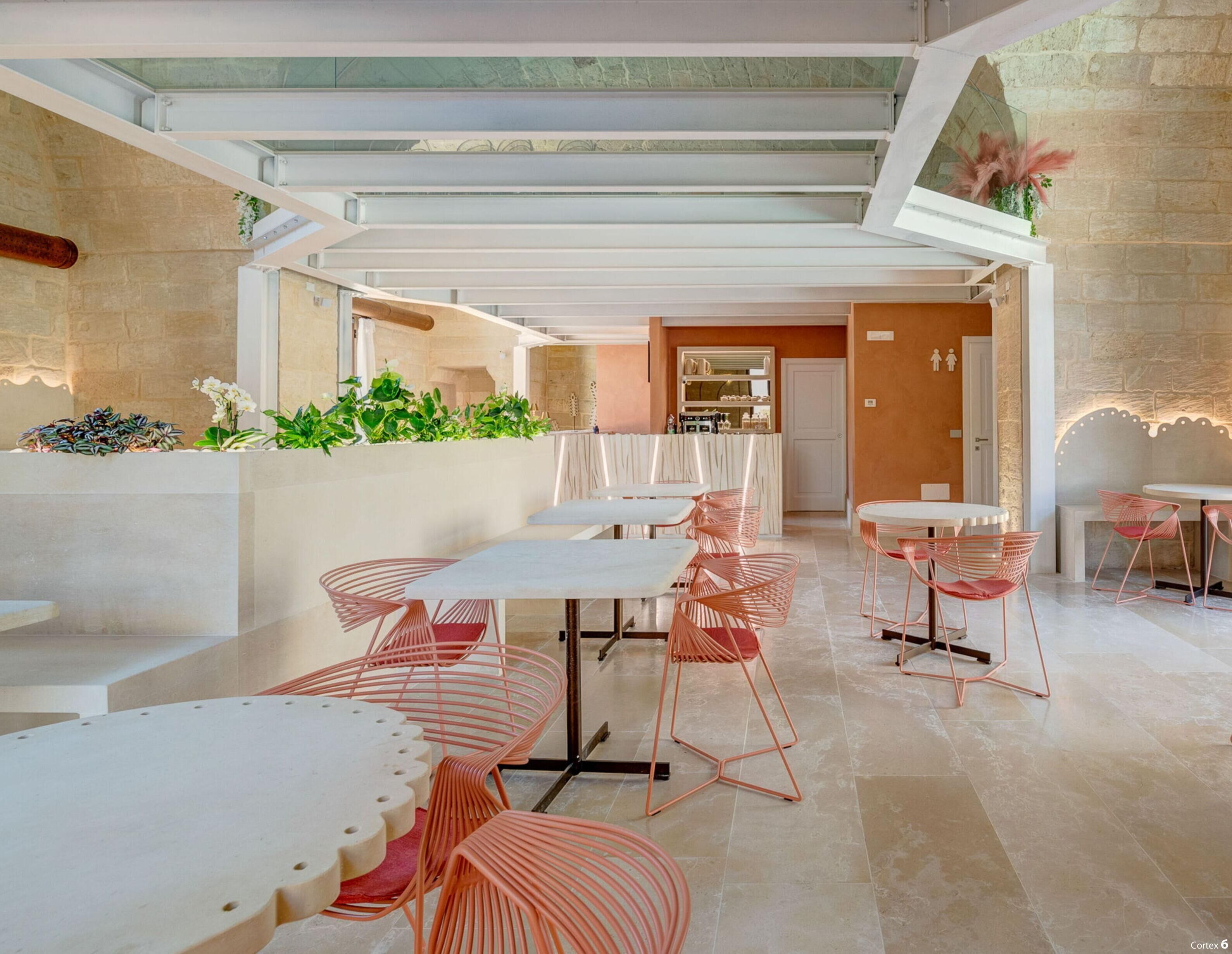 A modern restaurant with white tables and pink wire chairs, a stone wall, and a glass ceiling.