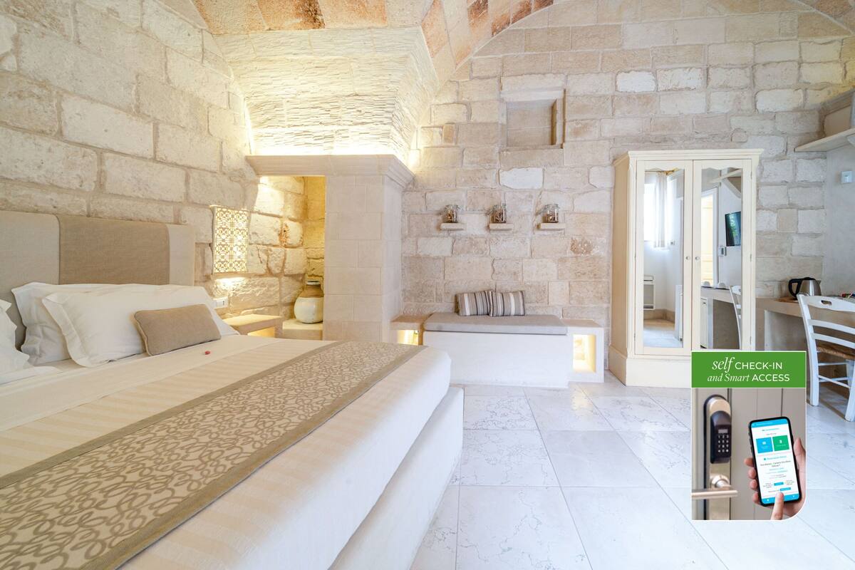 A modern hotel room with a stone wall, a bed with a patterned bedspread, a small seating area, and a self-check-in kiosk.