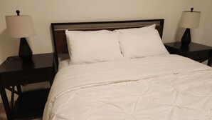 Un lit bien fait, recouvert de draps blancs, avec deux tables de chevet dotées de lampes, dans une chambre sobre agrémentée d’une tête de lit en bois.