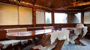 Une grande table à manger en bois, entourée de bancs, recouverte de plaids en fausse fourrure blanche, se trouve dans une pièce aux murs en bois et au sol carrelé.