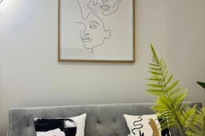 A framed line drawing of two faces on the wall, a grey sofa with patterned cushions, and a green plant on the right. - R&K Apartment Madrid - Comfortable and functional Standard Apartment in town (Madrid)