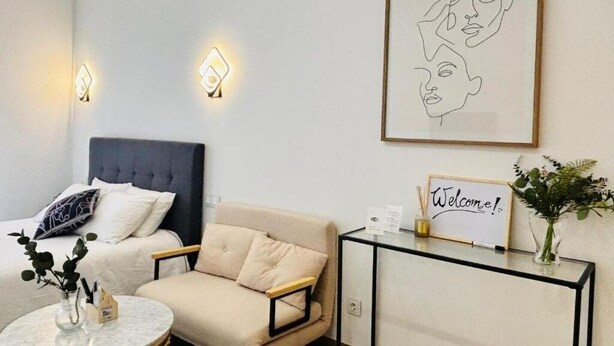 A modern hotel room with a bed, a sofa, a small table, and a welcome sign.