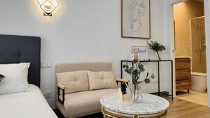 A hotel room with a bed, a sofa, a round table with a vase, a wall-mounted light, and a framed picture.