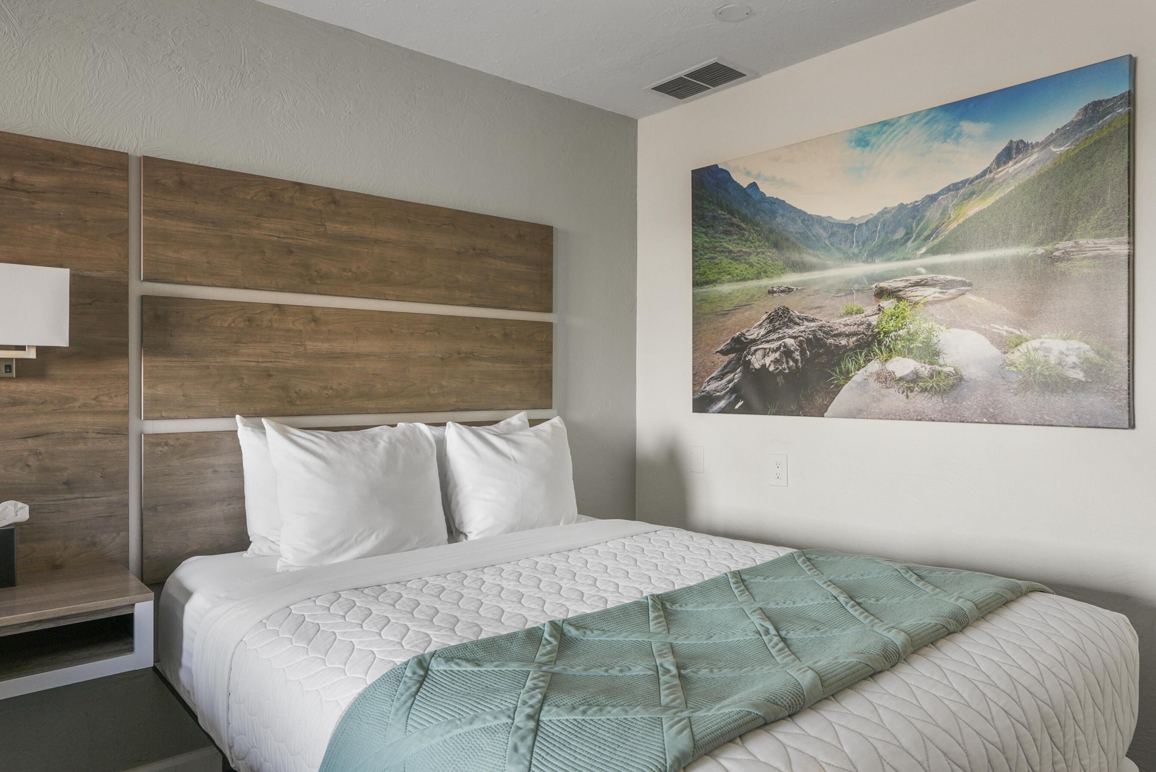 Een slaapkamer met een bed, een hoofdbord met een houten paneelontwerp, een schilderij van een bergachtig landschap en een nachtkastje met een lamp.
