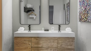 Ein Badezimmer mit weißem Waschtisch, zwei Spiegeln, einem Holzschrank und einem farbenfrohen, abstrakten Gemälde an der Wand.