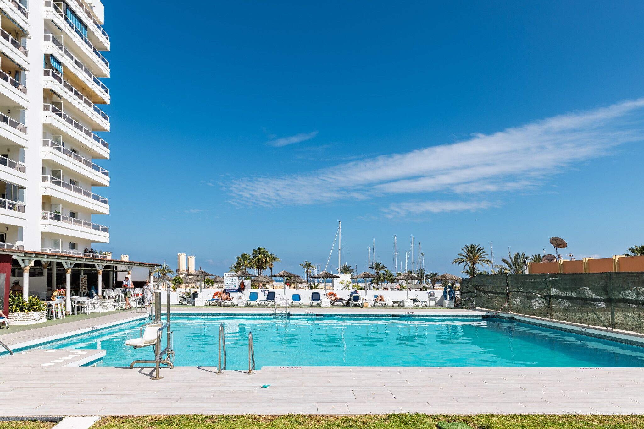 A swimming pool with lounge chairs and umbrellas, surrounded by a white building with multiple balconies.
