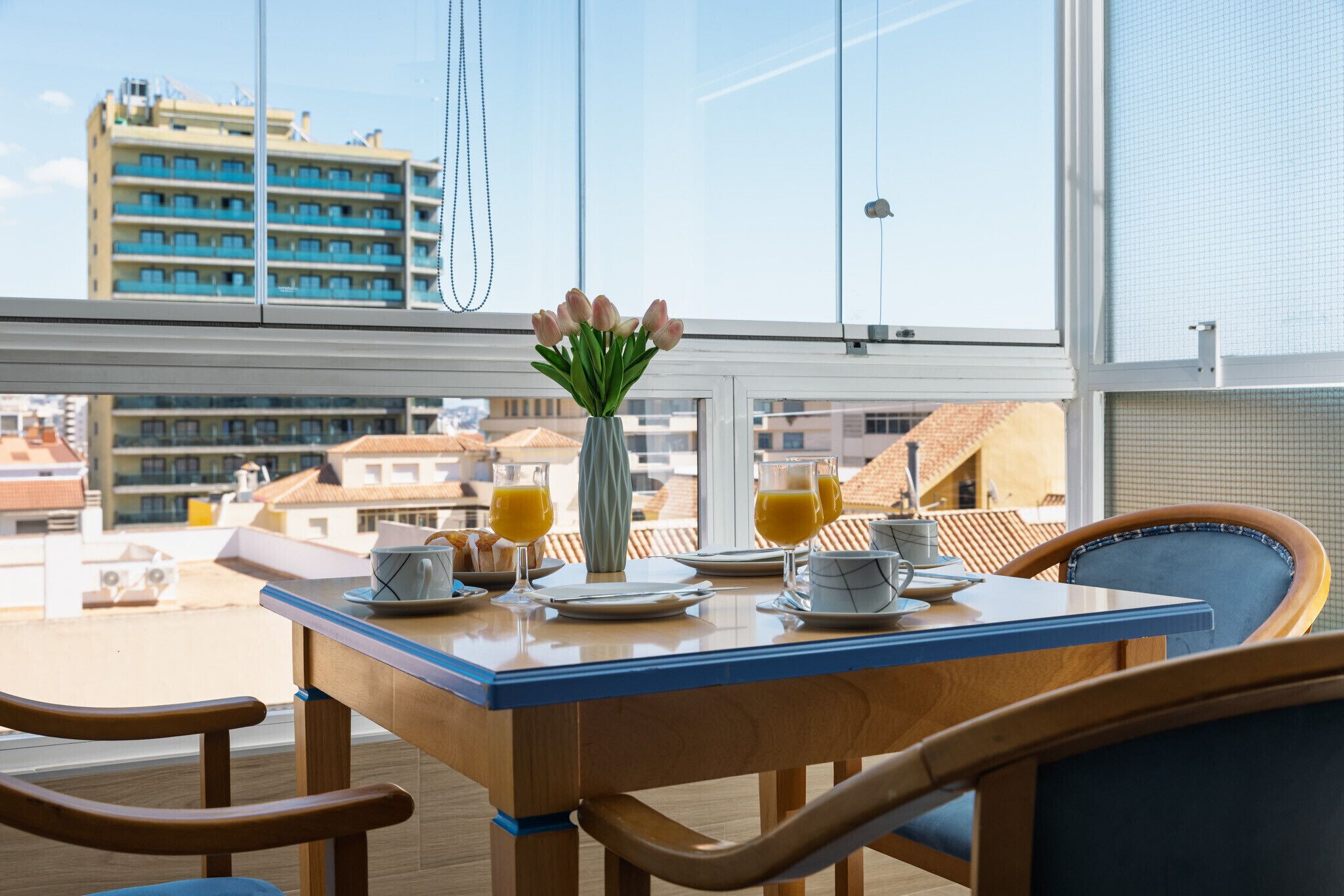 Une table à manger ornée d’un vase de fleurs, de deux verres de jus d’orange et d’une tasse, le tout placé sur un balcon donnant sur un paysage urbain.