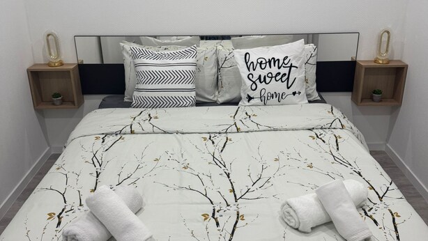A neatly made bed with a tree branch pattern bedspread, two pillows with black and white patterns, and a wall-mounted abstract art piece.