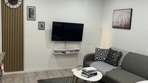 A modern living room with a flat-screen TV, a grey sofa, patterned cushions, a small round coffee table, and a decorative wall clock.
