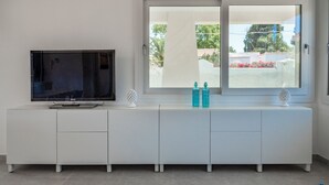 Een moderne woonkamer met een flatscreen-tv op een witte kast, een groot raam met uitzicht op groen en gebouwen, en twee decoratieve flessen op de kast.
